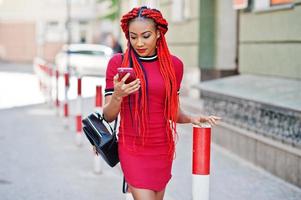 Cute and slim african american girl in red dress with dreadlocks and backpack posed outdoor and looking at mobile phone on street. Stylish black model. photo