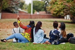 Back of group five african college students spending time together on campus at university yard. Black afro friends sitting on grass and studying with laptops. photo