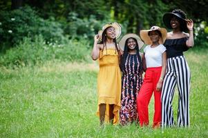 Group of four gorgeous african american womans wear summer hat spending time at green grass in park. photo