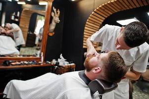 Young bearded man getting haircut by hairdresser while sitting in chair at barbershop. Barber soul. photo