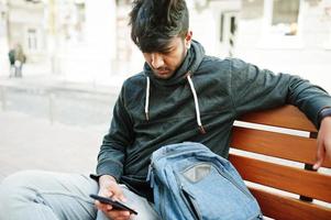 Portrait of young stylish indian man model pose in street, sitting on bench and hold smartphone at hand. photo