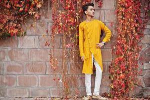 Indian stylish man in yellow traditional clothes posed outdoor against red leaves wall. photo