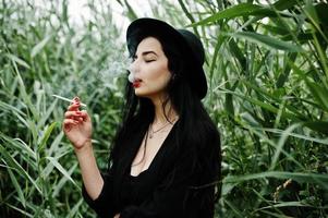 Sensual smoker girl all in black, red lips and hat. Goth dramatic woman smoking on common reed. photo