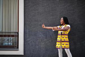 Stylish african american women in yellow jacket posed on street with hot drink in disposable paper cup and mobile phone at hands. photo