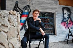 Young stylish macho boy in black jacket posed outdoor of street. Amazing model man sitting against grafiti wall of barbershop. photo