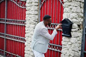 Rich and stylish african american man in blazer and eyeglasses check his mailbox outdoor in fence. photo