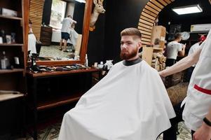 Young bearded man getting haircut by hairdresser while sitting in chair at barbershop. Barber soul. photo