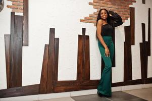 Beauty slim african american model wear on black blouse and green long legs pants posed against wooden wall at pub. photo