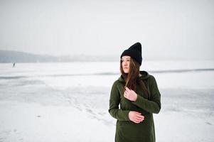 Funny girl wear on long green sweatshirt, jeans and black headwear, at frozen lake in winter day. photo