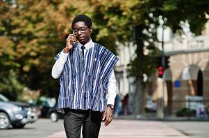 hombre africano con ropa tradicional y anteojos caminando en el cruce de peatones y hablando por teléfono móvil. foto