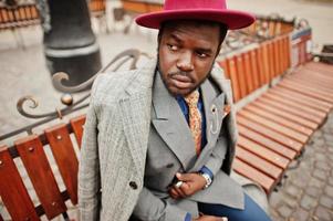 Stylish African American man model in gray coat, jacket tie and red hat posed at bench. photo