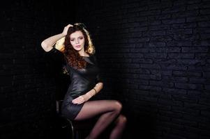 chica morena de pelo rizado y piernas largas con vestido de cuero negro posó en el estudio en una silla contra una pared de ladrillo oscuro. foto
