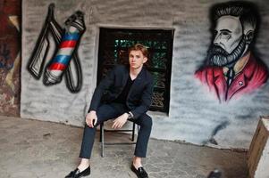 Young stylish macho boy in black jacket posed outdoor of street. Amazing model man sitting against grafiti wall of barbershop. photo
