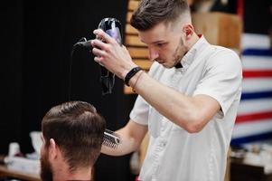 Young bearded man getting haircut by hairdresser while sitting in chair at barbershop. Barber soul. photo