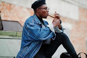 African american man in jeans jacket, beret and eyeglasses, lights a cigar and posed against btr military armored vehicle. photo