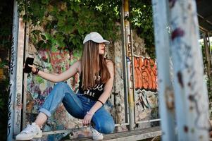 Stylish casual hipster girl in cap and jeans wear, listening music from headphones of mobile phone against large graffiti wall. photo