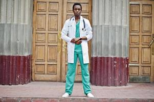 Stylish african american doctor with stethoscope and lab coat posed against door of hospital. photo