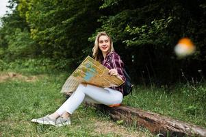 Portrait of a positive young gorgeous blonde sitting on the ground with a map in her hands in the forest. photo