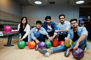 Group of five south asian peoples having rest and fun at bowling club. Holding bowling balls and sitting on alley. photo