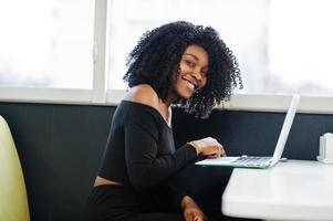 joven y bella mujer de negocios afroamericana de moda con peinado afro vestido de negro elegante, sentada y trabajando en una laptop. foto