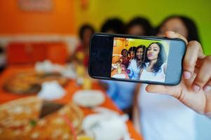 Close up photo phone screen of four young african girls in bright colored restaurant eating pizza, having fun together and making selfie.