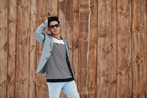 Casual young indian man in silver blazer and sunglasses posed against wooden background. photo