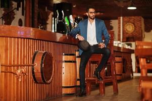 Handsome well-dressed arabian man with glass of whiskey and cigar posed at pub. photo