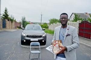 Rich and stylish african american man in blazer and white pants, eyeglasses read magazine against his suv car. photo