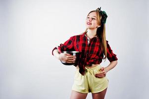 Young funny housewife in checkered shirt and yellow shorts pin up style with saucepan and kitchen spoon isolated on white background. photo