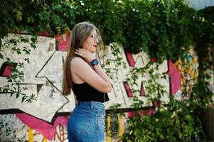 Stylish casual hipster girl in cap, glasses and jeans wear, listening music from headphones of mobile phone against large graffiti wall. photo