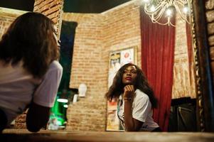 Stylish african american woman in printed t-shirt looking at mirror. photo