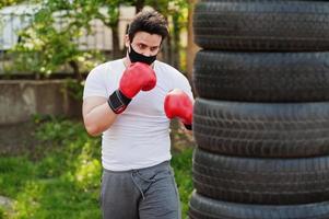 retrato deportivo boxeador árabe con mascarilla médica negra boxeando al aire libre durante la cuarentena del coronavirus. foto