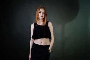 Portrait of a beautiful redheaded girl in black top and black skirt posing in the studio next to the grey wall. photo