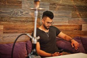 Stylish beard arabian man in glasses and black t-shirt smoking hookah indoor bar. Arab model having rest and looking at phone. photo