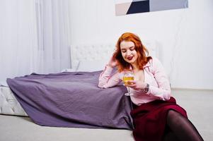 Gorgeous red haired girl in pink blouse and red skirt with glass of wine at hand near bed at room. photo