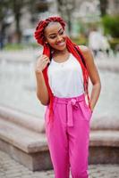 chica afroamericana de moda en pantalones rosas y rastas rojas posadas al aire libre contra fuentes. foto