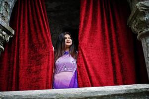 niña hindú india en el sari violeta tradicional posó en la calle contra la casa antigua con cortinas rojas. foto