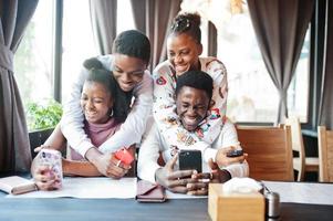 Happy african friends sitting and chatting in cafe. Group of black peoples meeting in restaurant hugs and look at their mobile phone. photo