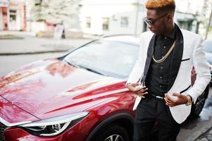 hombre afroamericano elegante y guapo con traje blanco contra un coche de lujo rojo. foto