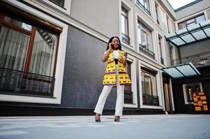 Stylish african american women in yellow jacket posed on street with hot drink in disposable paper cup and mobile phone at hands. photo