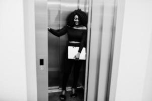 moda joven hermosa mujer de negocios afroamericana con peinado afro en elegante negro, de pie en el ascensor con el portátil en las manos. foto