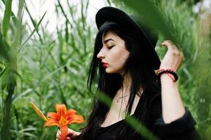 chica sensual todo en negro, labios rojos y sombrero. mujer dramática gótica sostiene flor de lirio naranja en caña común. foto