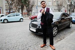 Stylish indian businessman in formal wear tied his tie and standing against black business car on street of city. photo