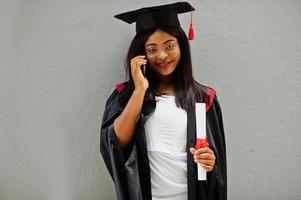 joven estudiante afroamericana con diploma y teléfono móvil posa al aire libre. foto