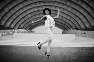 Increíble mujer modelo afroamericana con pantalones verdes y sombrero negro posada al aire libre contra el salón de la arena, salta en el aire. foto
