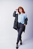 Portrait of a fantastic redheaded girl in blue shirt, grey overcoat posing with sunglasses in the studio. photo