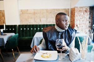 hombre africano con ropa tradicional negra sentado en el restaurante y comiendo pasta y bebiendo vino. foto