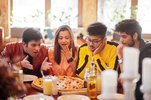 grupo de amigos asiáticos comiendo pizza durante la fiesta en la pizzería. gente india feliz divirtiéndose juntos, comiendo comida italiana y sentados en el sofá. caras de asombro y sorpresa. foto