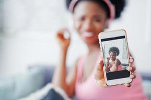 foto de la pantalla del teléfono móvil joven afroamericana sentada en el sofá, escuchando música en los auriculares.