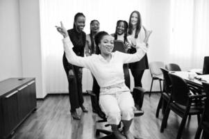 Five multiracial business womans standing at office and roll woman on chair. Diverse group of female employees in formal wear having fun. photo